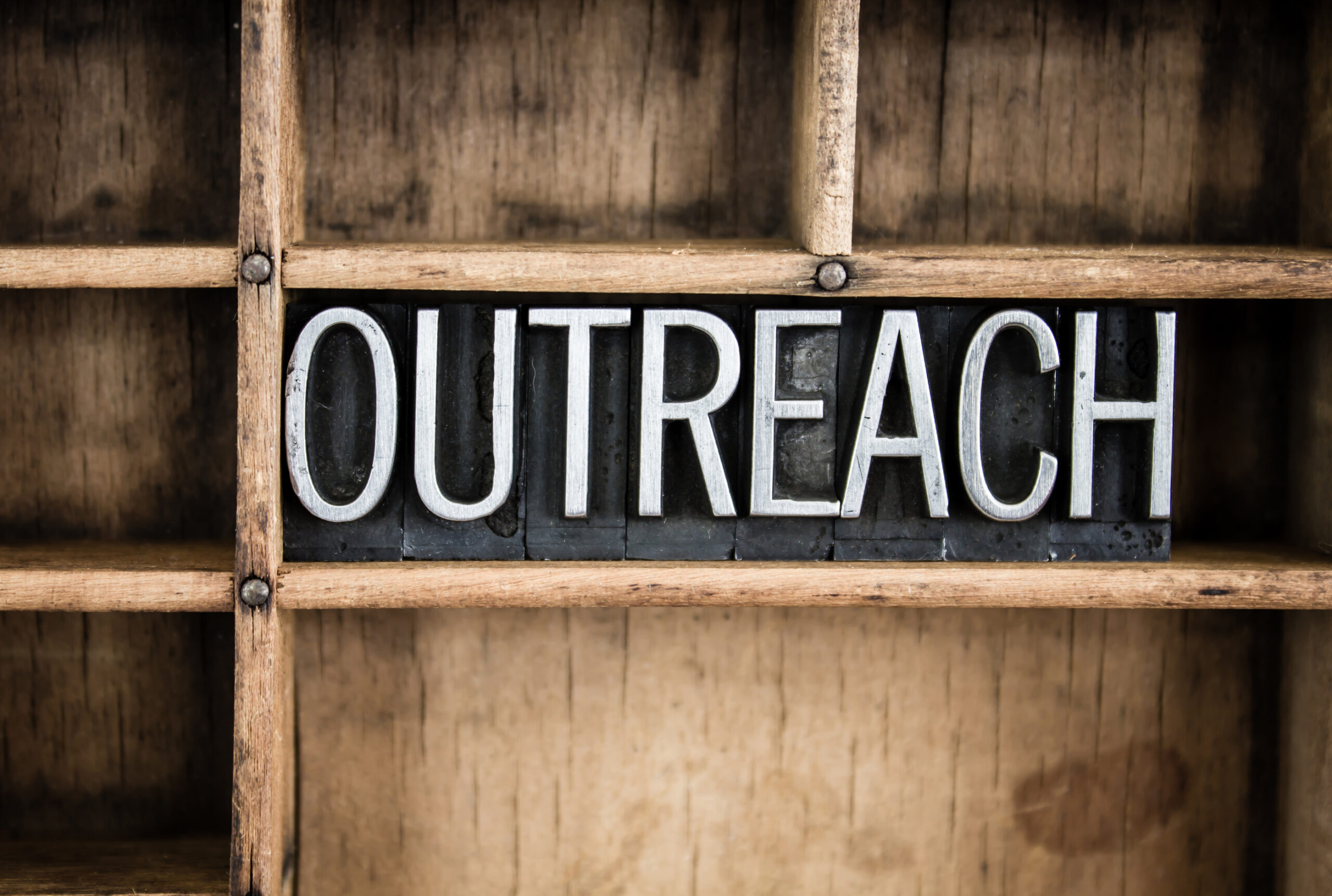 The word "OUTREACH" written in vintage metal letterpress type in a wooden drawer with dividers.