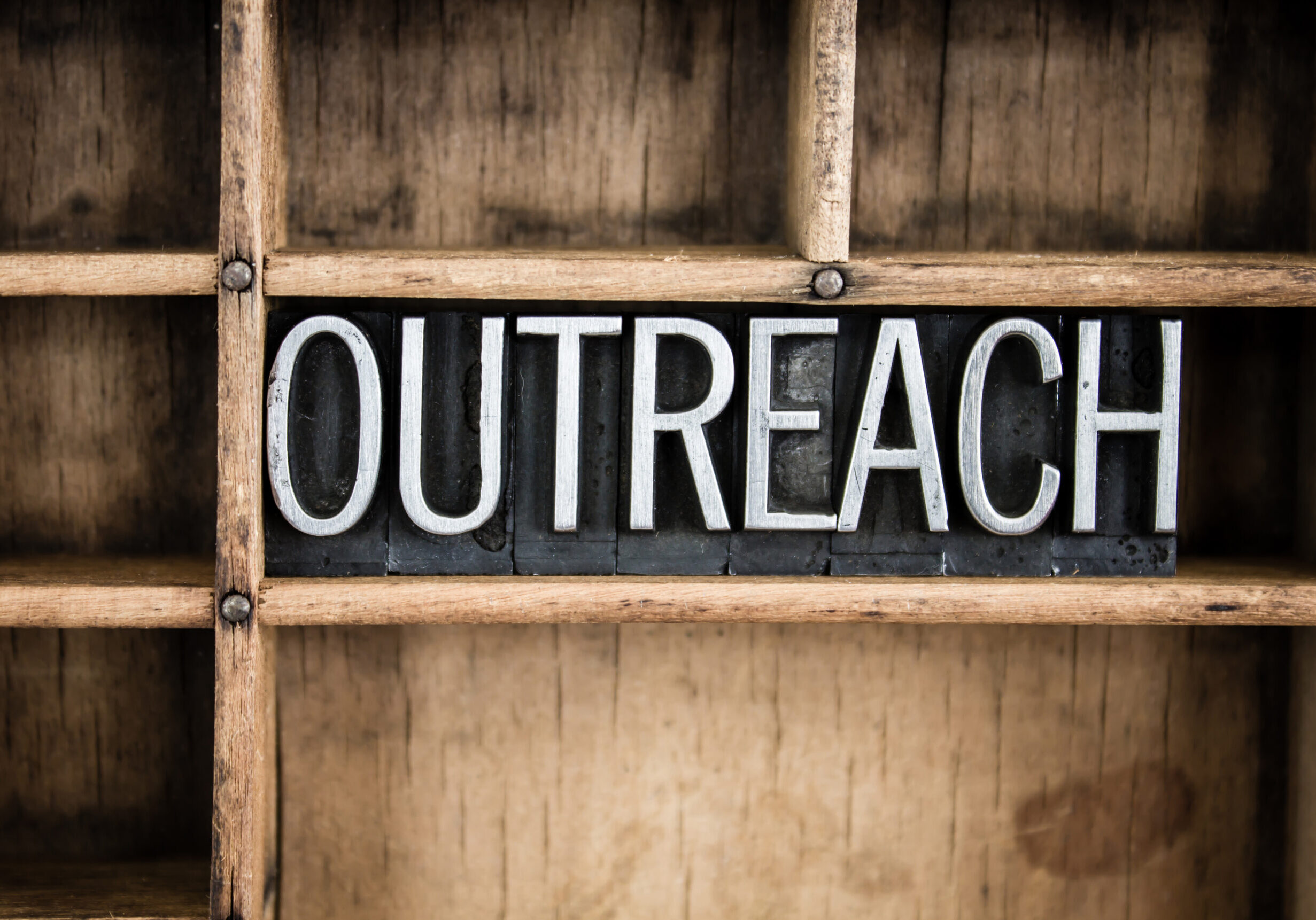 The word "OUTREACH" written in vintage metal letterpress type in a wooden drawer with dividers.